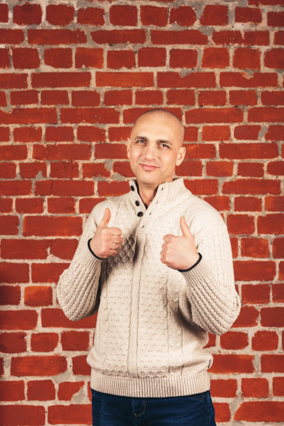 穿着白毛衣的秃子在砖墙背景上竖起大拇指。副本的空间。图片下载