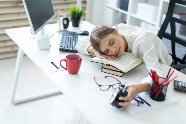 一个年轻的女孩正坐在办公室的桌子旁，把头靠在桌子上，手里握着闹钟。图片下载