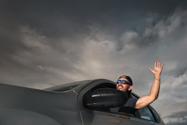 戴着太阳镜的微笑男司机。他举起了手。正在下雨，阳光灿烂图片下载