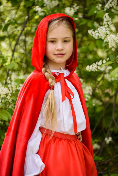 微笑的金发女孩穿着小红帽在绿树旁摆姿势图片下载