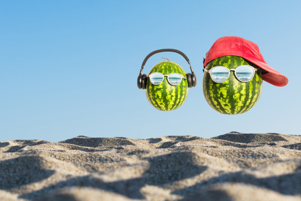 这是一幅夏天的创意照片，在沙滩上的玻璃杯里有两个脑袋形状的西瓜。图片下载
