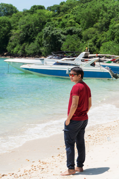 一名亚洲男子站在海滩上，以快艇为背景望着大海。图片下载