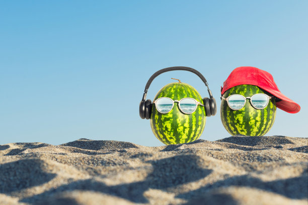 这是一幅夏天的创意照片，在沙滩上的玻璃杯里有两个脑袋形状的西瓜。图片下载