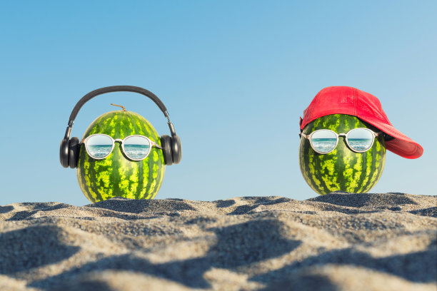 这是一幅夏天的创意照片，在沙滩上的玻璃杯里有两个脑袋形状的西瓜。图片下载