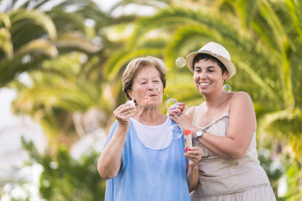 母女白种人夫妇一起玩吹肥皂泡在户外休闲活动。自然绿色背景散焦。快乐和顽皮的人们在友谊的肖像图片下载