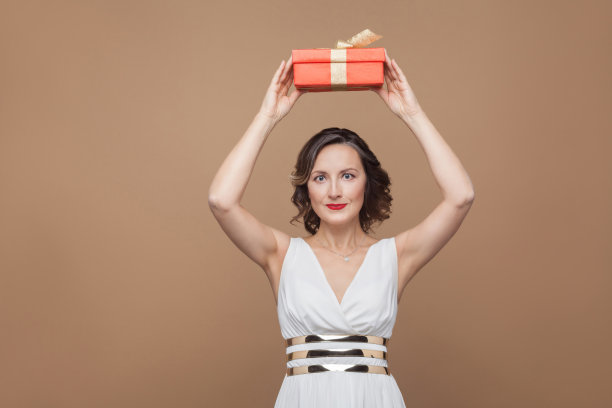 美丽的黑发女人在白色的裙子拿着红色的礼品盒在头顶和看着镜头微笑。情感和感觉的概念。图片下载