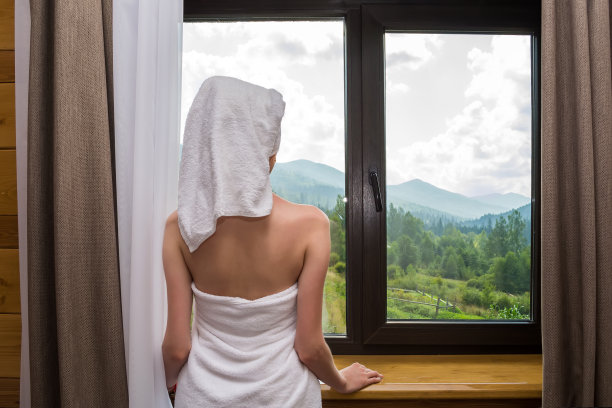 一个年轻、美丽、性感的女人，洗完澡后，裹着毛巾站在靠近酒店窗户的地方，可以看到群山图片下载