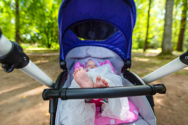 可爱的小女婴坐在婴儿车里，双腿搭在婴儿车的扶手上。父母带着孩子在公园里散步。图片下载