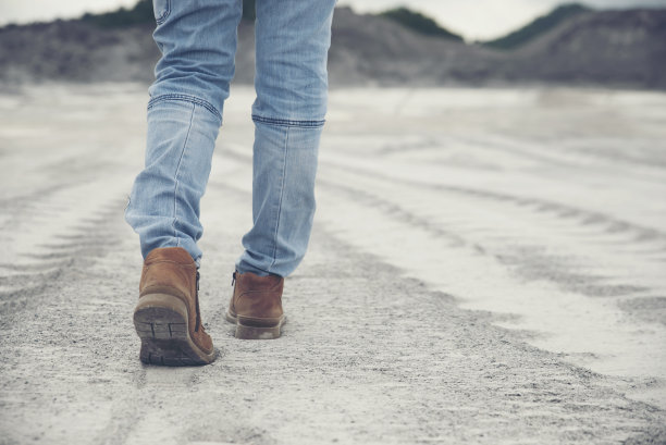 人的后视图(孤独女孩)脚的图片，穿着棕色皮靴和蓝色牛仔裤。旅行者的脚步走向成功的小路和绿色的田野旁边。旅行与成功的概念图片下载