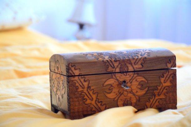 一个装饰过的小木箱放在一张铺着橙色床上的床上，背景里有几个枕头，白色的墙壁和窗帘旁边还有一盏白色的灯。素材图片