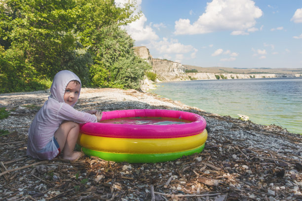 小男孩坐在海边，在充气游泳池里玩耍，背景是如画的风景。图片下载
