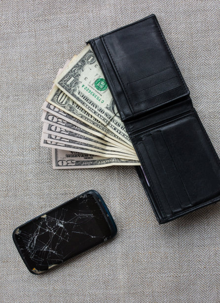 崩溃的黑色智能手机和钱包里的钱买一个新的。质朴的桌布上放着美元和破手机。是时候为电子产品贷款了。垂直的照片。图片下载