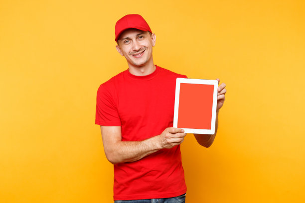 快递员被隔离在桔黄色背景上。职业男员工快递员，红帽，t恤，手持平板电脑与空白空白屏幕。服务的概念。副本的空间。模型模板。图片下载