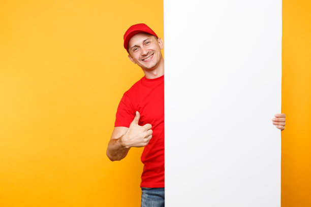身穿红色制服的送货员被隔离在黄橙色背景下。男员工快递员戴着便帽，t恤上拿着大大的空白空白广告牌与文案空间，放置文字图像。服务的概念。图片下载