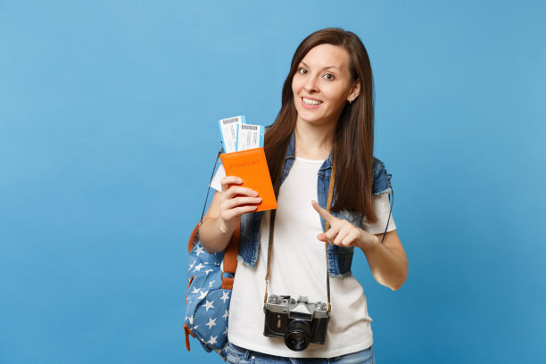 年轻漂亮的女学生与复古照片相机颈部指向的食指在护照，登机牌票孤立在蓝色背景。在国外的大学教育。航空飞行。图片下载