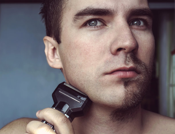 年轻的大胡子男子在使用修剪器修剪胡须。半张脸，半张胡子图片下载