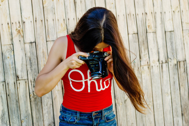 年轻美丽的黑发女孩在红色衬衫和牛仔裤短裤摆姿势与相机在乡村木制背景。拍照片，拍照片，女人，摄影师，老式相机，户外人像图片下载