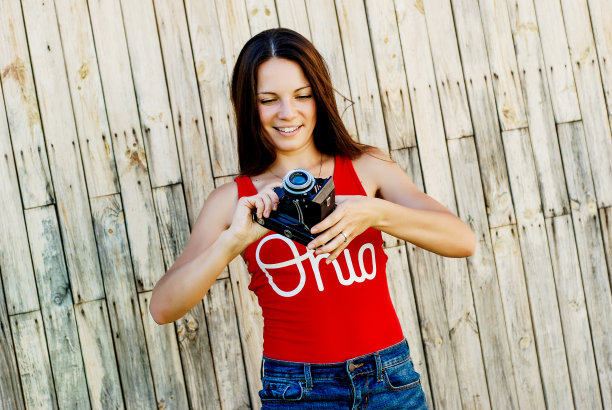 年轻美丽的黑发女孩在红色衬衫和牛仔裤短裤摆姿势与相机在乡村木制背景。拍照片，拍照片，女人，摄影师，老式相机，户外人像图片下载