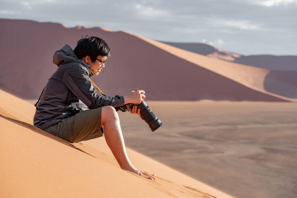 年轻的亚洲男子旅行者和摄影师坐在纳米比亚沙漠的沙丘上看相机上的照片显示，非洲纳米比亚。旅行摄影的概念图片下载