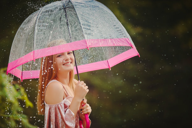 一个十几岁的女孩在穿过公园时躲在伞下避雨。她大笑，微笑，享受温暖的雨天。图片下载