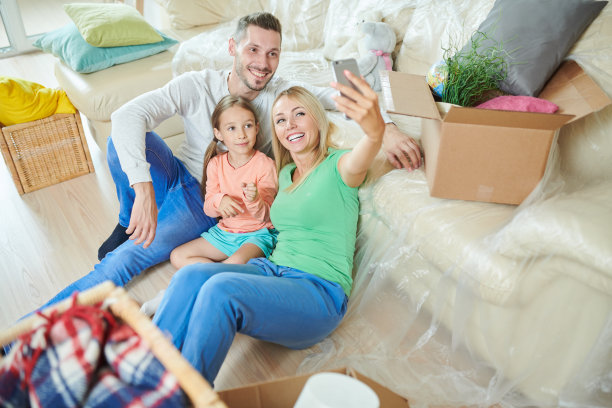 欢乐的年轻家庭穿着休闲服装坐在地板上，倚在沙发上自拍进入新家，沙发和地板上满是盒子图片下载
