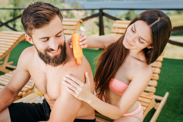 这是一张漂亮的照片，细心的女孩坐在男孩后面，从她手里拿着的橙色瓶子里拿出一些防晒霜。女人揉了揉他的肩膀。大胡子向后看，笑了图片下载