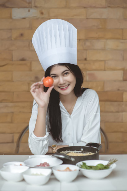 漂亮的年轻女子戴着厨师帽，正在做炒辣意大利面，看着镜头微笑着在家厨房烘焙，也被称为醉意大利面或Phad Kee Mao意大利面图片下载