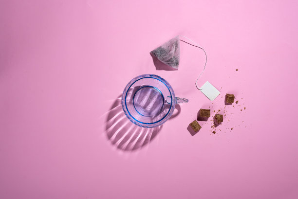 这是一张创意的照片，上面是一个蓝色的玻璃杯，有着美丽的反光阴影，粉红色的背景上是一袋茶和红糖，俯视图图片下载