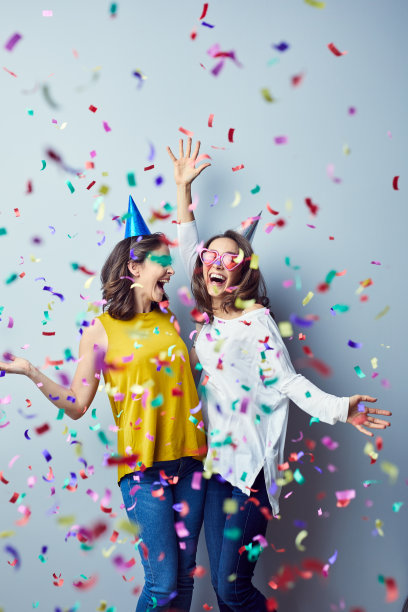 一对漂亮的年轻女性大笑着举起手臂，用生日帽和五彩纸屑饰品来欢呼图片下载