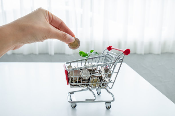 女人用手把硬币放进玩具购物车和绿色植物里。省钱的概念是购物。图片下载