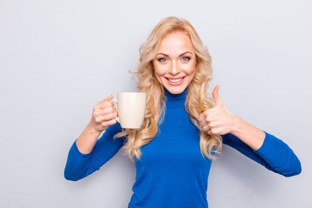 漂亮的，时髦的，迷人的，漂亮的，可爱的女人在蓝色的翻领孤立在灰色的背景有杯子和茶在手里显示拇指向上与手指看着相机图片下载