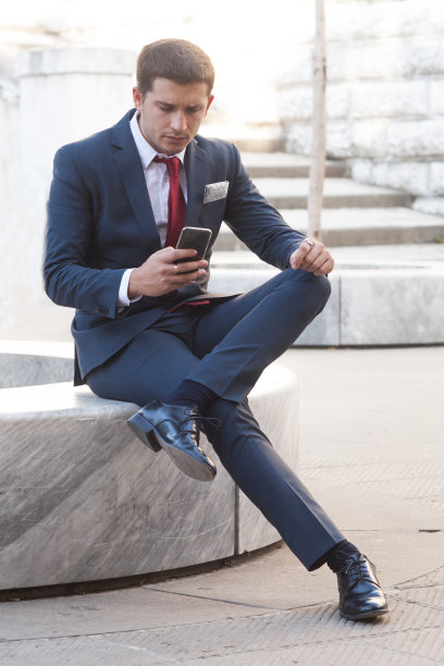 年轻的商人穿着西装打着领带坐在公园的大理石长凳上看智能手机图片下载