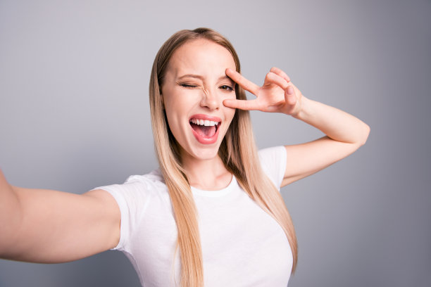 年轻的白人迷人迷人的微笑女孩的肖像显示v-sign和平手势在摄像头自拍照片。在灰色背景上隔离图片下载