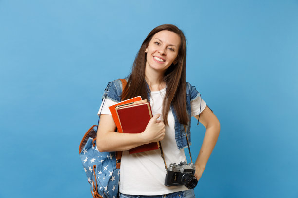 年轻微笑的女学生的肖像背包和复古的照片相机在脖子上拿着学校的书孤立在蓝色的背景。中学教育中的大学学院观念。图片下载