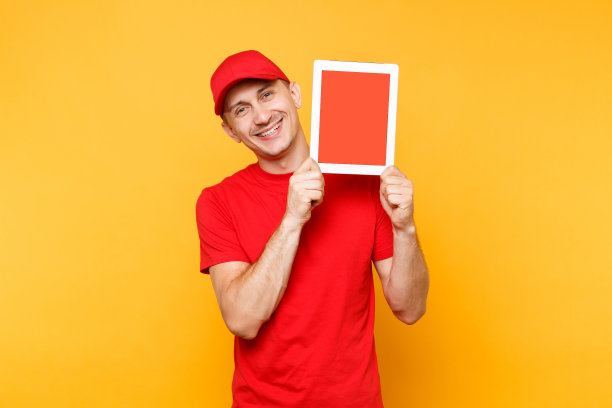 快递员被隔离在桔黄色背景上。职业男员工快递员，红帽，t恤，手持平板电脑与空白空白屏幕。服务的概念。副本的空间。模型模板。图片下载