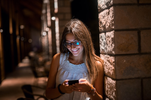 美丽的年轻女孩靠在墙上看着她的手机。微笑着，充满爱意。图片下载