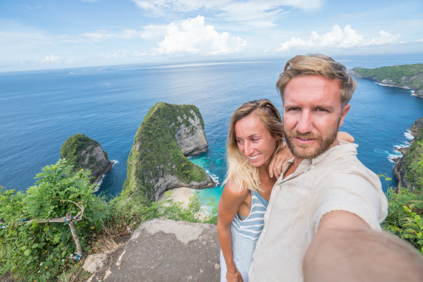 一对年轻夫妇在克林金海滩上自拍——蓝色和绿色的沙子，美丽的山在海上形成。旅游目的地度假的人喜欢分享社交媒体概念的自然风景图片下载