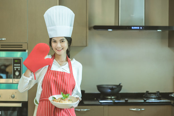 漂亮的年轻女子戴着厨师帽，正在做炒辣意大利面，看着镜头微笑着在家厨房烘焙，也被称为醉意大利面或Phad Kee Mao意大利面图片下载