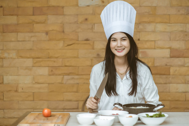 漂亮的年轻女子戴着厨师帽，正在做炒辣意大利面，看着镜头微笑着在家厨房烘焙，也被称为醉意大利面或Phad Kee Mao意大利面图片下载