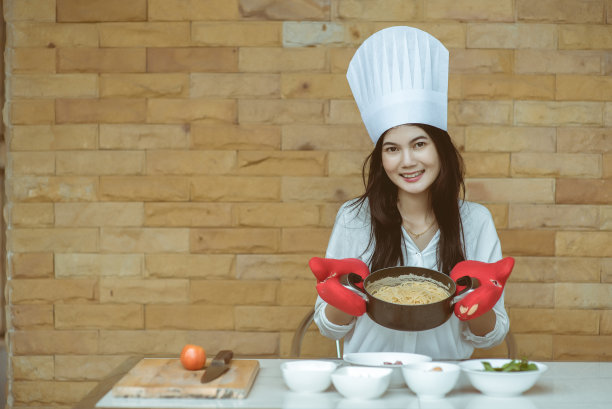 漂亮的年轻女子戴着厨师帽，正在做炒辣意大利面，看着镜头微笑着在家厨房烘焙，也被称为醉意大利面或Phad Kee Mao意大利面图片下载