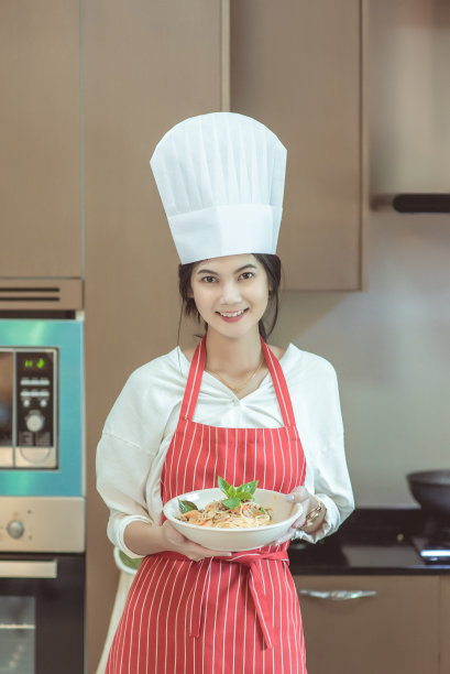 漂亮的年轻女子戴着厨师帽，正在做炒辣意大利面，看着镜头微笑着在家厨房烘焙，也被称为醉意大利面或Phad Kee Mao意大利面图片下载