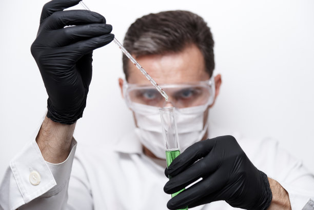 一个科学家化学家手里拿着一个试管，里面有一种绿色的液体，试管被隔离在白色的背景上。实验是由一位戴着护目镜、口罩和手套的科学家进行的。图片下载