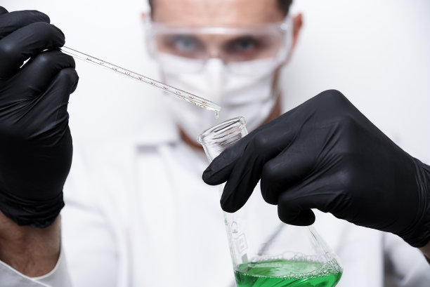 在白色背景下，戴着防护眼镜、面罩和黑手套的科学家手中，一根试管从玻璃烧瓶中掉落。图片下载
