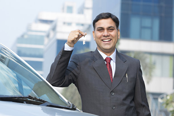 穿着西装，手里拿着车钥匙的成年男性商人。汽车在背景-库存图像图片下载