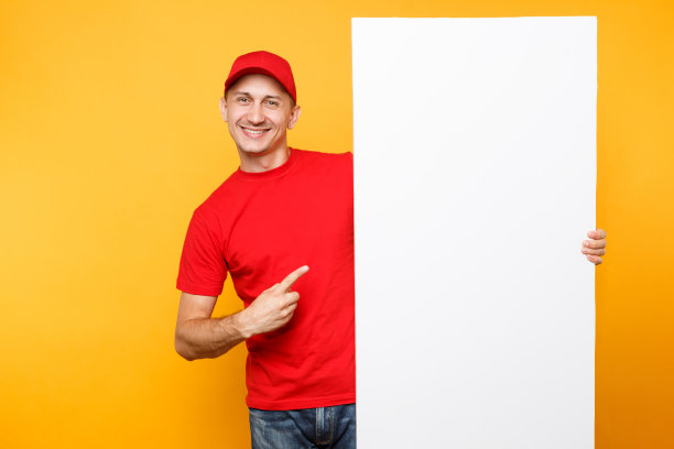 身穿红色制服的送货员被隔离在黄橙色背景下。男员工快递员戴着便帽，t恤上拿着大大的空白空白广告牌与文案空间，放置文字图像。服务的概念。图片下载