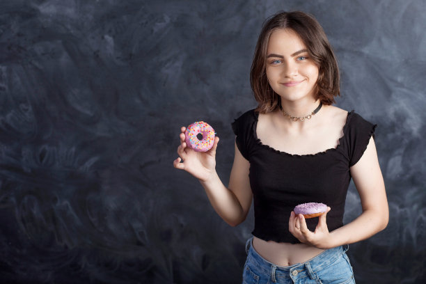 快乐女孩的肖像与油炸圈饼在黑色的背景。快乐的女孩拿着新鲜的甜甜圈微笑着。好心情，饮食观念。图片下载
