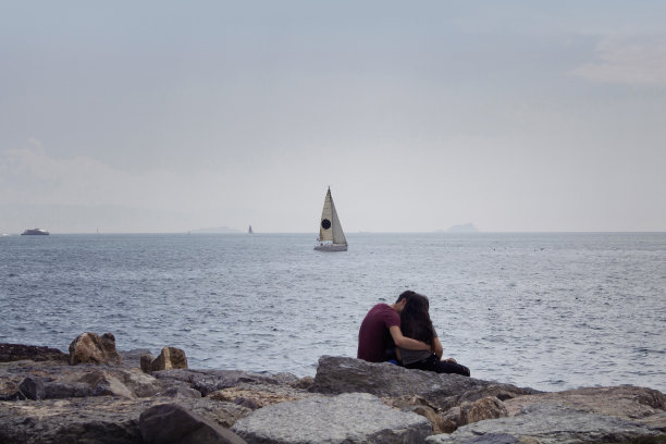 一对陌生的年轻夫妇坐在博斯普鲁斯海岸边的岩石上。图片拍摄于伊斯坦布尔亚洲一侧名为Moda的社区。背景是一艘帆船。图片下载