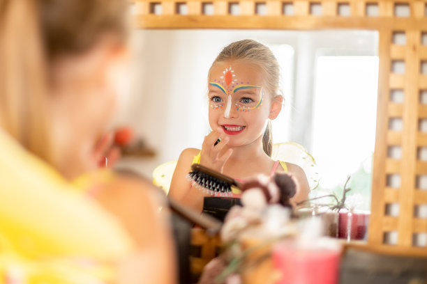 快乐的孩子带着脸上的彩绘，穿着万圣节的服装看着镜子，微笑着。兴奋的小女孩看着镜子里的自己。图片下载