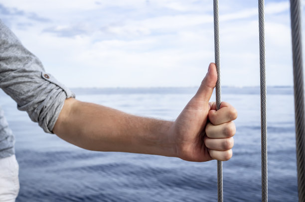 在这夏日的大海、海浪和云中天空的背景下，这名男子用大拇指向上握住游艇上的金属家伙的手。图片下载