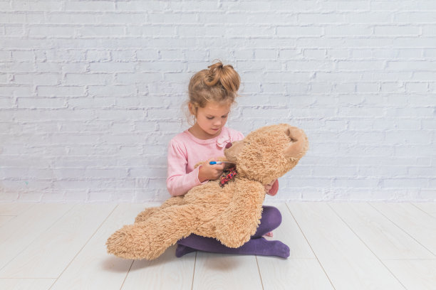 女孩，孩子测量一个玩具熊的温度，靠在一堵白色的砖墙图片下载